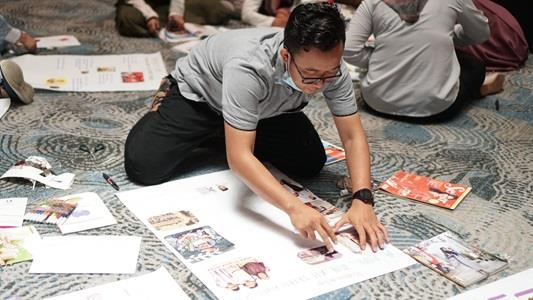 Asian an with glasses working with pen and paper on the floor, behind him other people